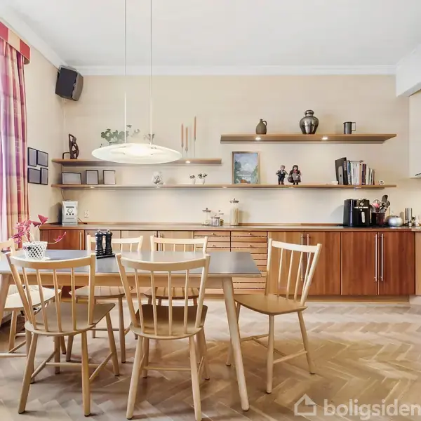 Et spisebord med seks stole står under en moderne lampe. Køkkenet har træskabe, åbne hylder med dekorationer og en ovn. Store vinduer med gardiner lyser rummet op.