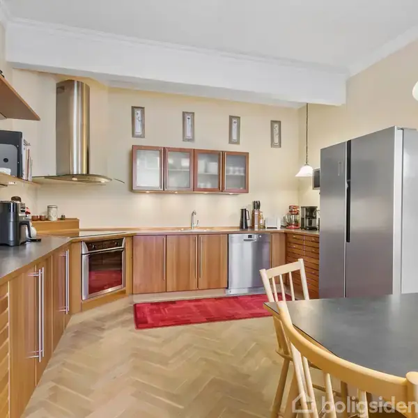 Køkken med træskabe, sort bordplade og rustfri stålhvidevarer. Kaffemaskine og køkkenredskaber står på bordet. Spisebord med stole i forgrunden, rød løber på gulvet.