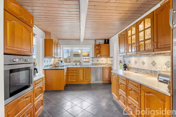 Køkken med træskabe og granitbordplader strækker sig langs væggene. Ovnen er integreret i venstre skabsvæg. Stort vindue over vasken giver udsigt til baghaven.