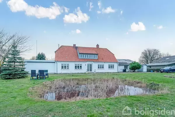 En hvid etplansvilla med rødt tegltag står foran en græsplæne med en lille dam. Der er nogle stole og et bord til venstre. Skyer dækker den blå himmel.