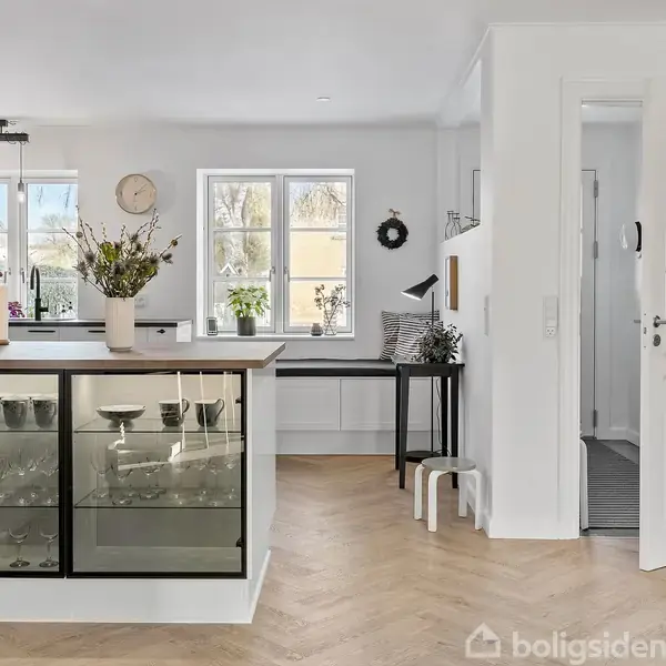 Glasvitrine oplyst indefra viser glasgenstande, placeret i et lyst køkken med træparketgulv. En vase med blomster står ovenpå, og et ur hænger på væggen nær vinduet.