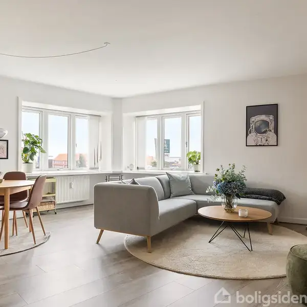 En lys stue med en grå sofa og runde sofabord, blomster i vase, og vægkunst. Spisebord med stole i baggrunden, ved store vinduer. Indrettet med moderne stil.