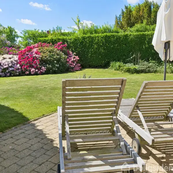 To træliggestole står på en terrasse, vendt mod en grøn have med blomstrende buske. En parasol står ved siden af stolene under en klar blå himmel.
