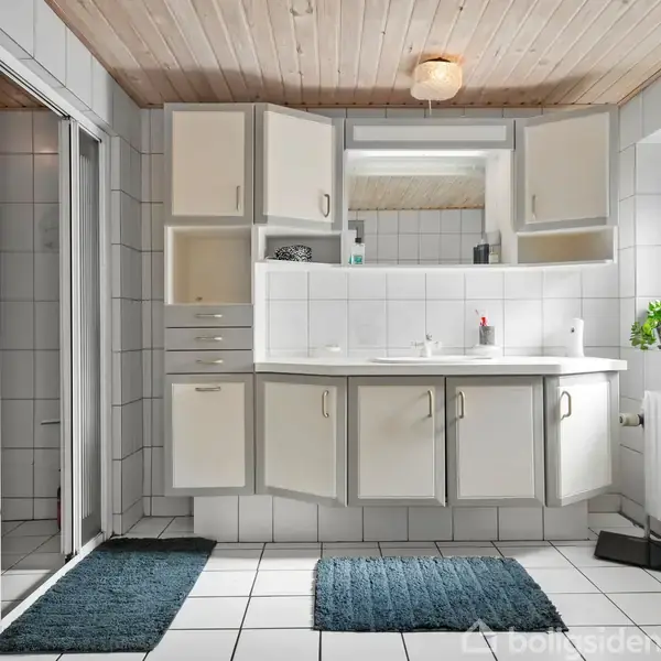 Et badeværelse med et hvidt skabsarrangement under en stor spejlskabsvæg. To tæpper ligger på det hvide flisegulv, og der er en plante ved vinduet.