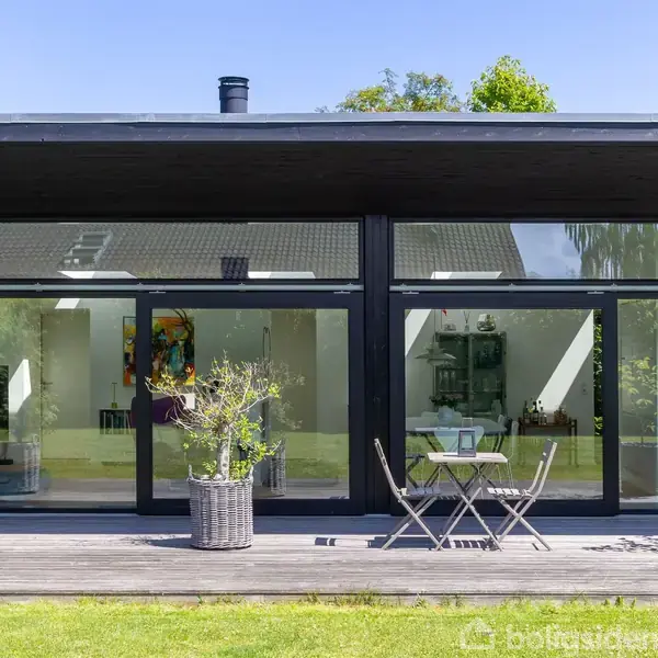 Et moderne hus med store glaspartier står i en have. Foran huset er der en træterrasse med et lille bord og to stole. Hele omgivelserne er grønne og frodige.