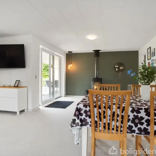 Spisebord med mønstret dug og træstole står nær brændeovn i moderne, lyst rum. Der er fladskærm på væggen og dobbeltglasdøre mod terrasse.