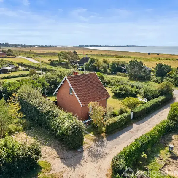 Rødt hus med skråt tag står omgivet af tæt grønt hegn og buske, ved en grussti i et naturskønt område tæt på kysten. Udsigt over havet i baggrunden.