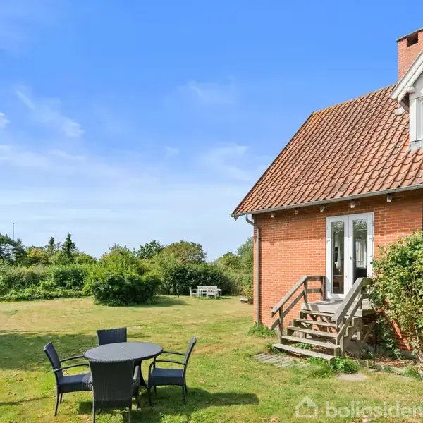 Et rødt murstenshus med trætage står på en græsplæne. Foran huset er der lukkede sorte havemøbler. Området er grønt med buske og et enkelt træ i baggrunden.