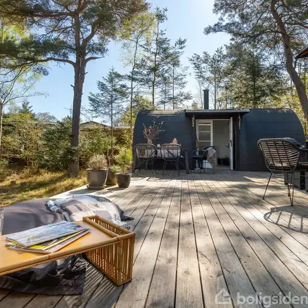 Terrasse med træplanker foran et moderne sommerhus. Et lavt bord med en flaske og magasiner står på gulvet. Omkranset af træer giver stedet en rolig udendørs atmosfære.