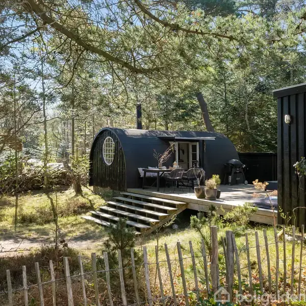 En sort, cylinderformet bygning med runde vinduer står på en træterrasse med trapper. Havemøbler er på terrassen. Omgivet af skovbevoksning og indhegnet med et lavt hegn.