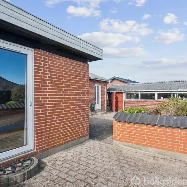 Røde murstenshuse med hvide vinduer og døre står i en udendørs gårdhave. Flisebelagt område med en lav mur og en planteboks. Skyer ses på en klar blå himmel.