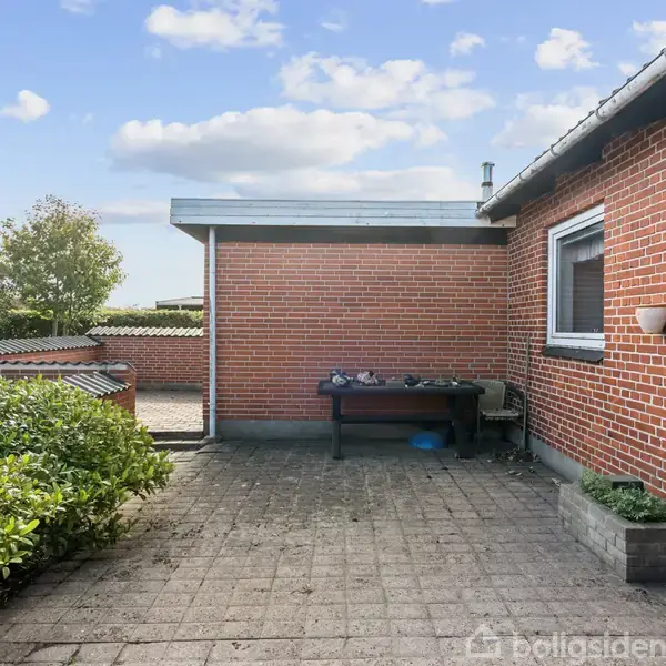 En rød murstensbygning med fastgjorte vinduer og en udendørslampe. Bygningen står omkring en flisebelagt terrasse med en bordfodbold, omkranset af en velholdt hæk under en blå himmel med skyer.