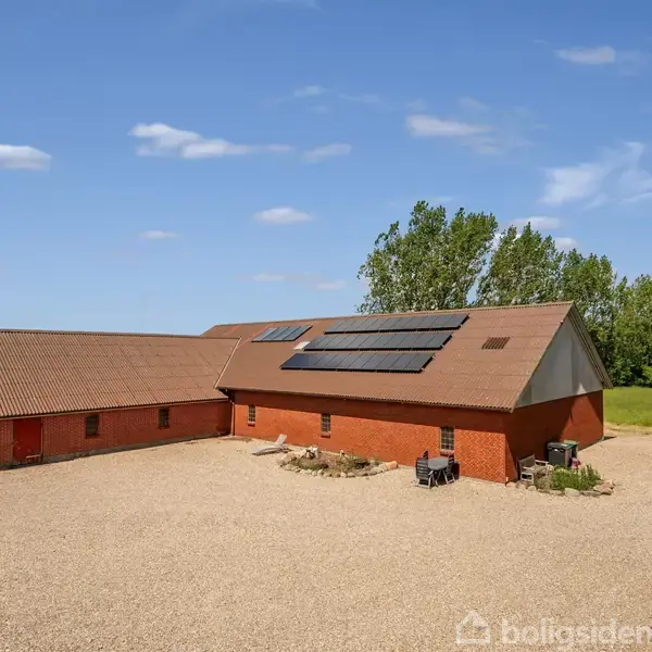 En rød murstensbygning med solpaneler på taget står stille på en gårdsplads med grus og omkranset af grønne træer under en blå himmel med lette skyer.