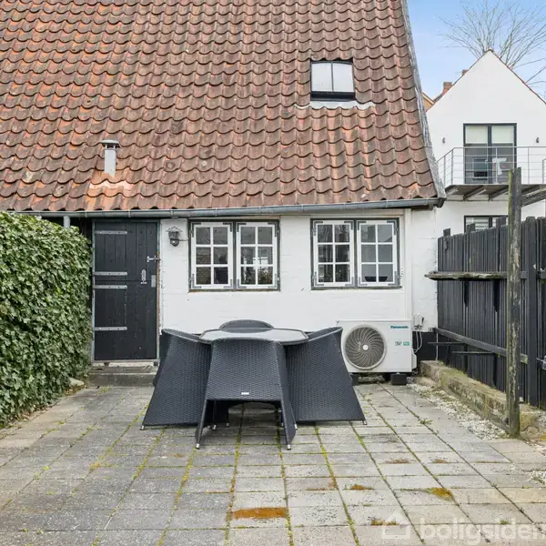 Et lille, hvidt hus med rødt tegltag omsluttet af hæk og hegn. En terrasse med fliser og et sort havemøbel sæt under åben himmel.