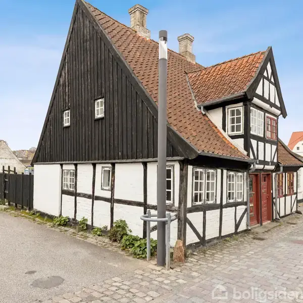 Et bindingsværkshus står stille med sort-hvid facade og rødt tegltag, placeret på brostensbelagt gade i en bymæssig miljø med flere omkringliggende bygninger.
