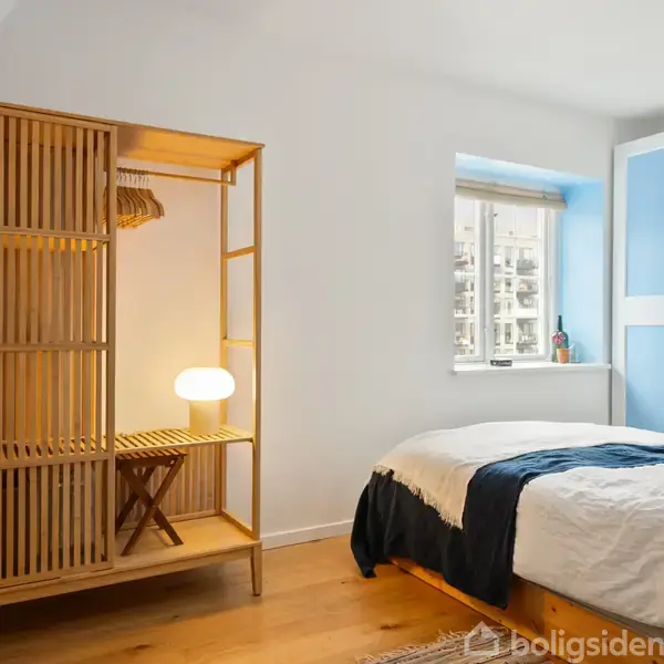 Trægarderobe står til venstre med en tændt lampe indeni. En redt seng er til højre. Værelse har trægulv og vindue med udsigt. Hvid og blå skabsdør i baggrunden.