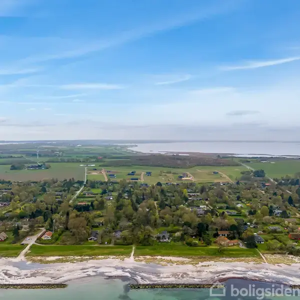 Kystområde med strande og bølger i forgrunden; bagved ses grønne marker og spredte huse, omgivet af frodig vegetation under en klar blå himmel.