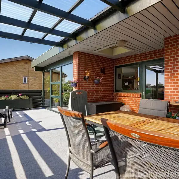 Terrasse med træbord og stole står under glasoverdækning. Omgivet af røde murstensvægge og vinduer, fremhæver blomsterkrukker hygge omkring. Naboens hus kan ses i baggrunden.