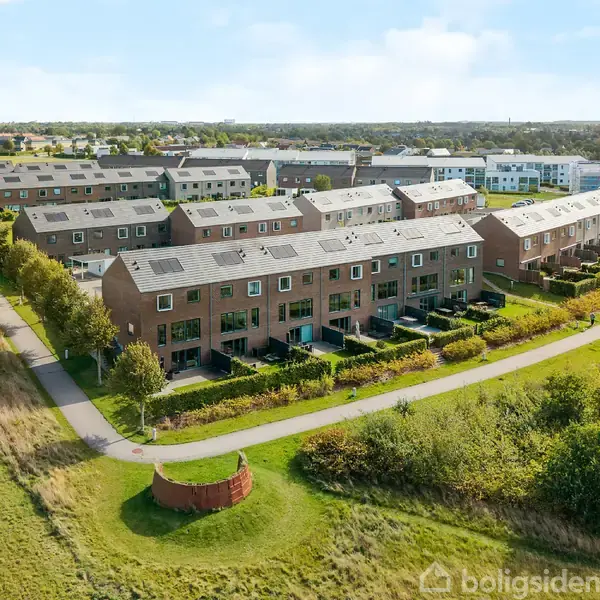 Rækkehuse i røde mursten står på række med solpaneler på taget, omgivet af stier og grønne områder i et forstadsområde. Du kan se en lang række af disse huse.