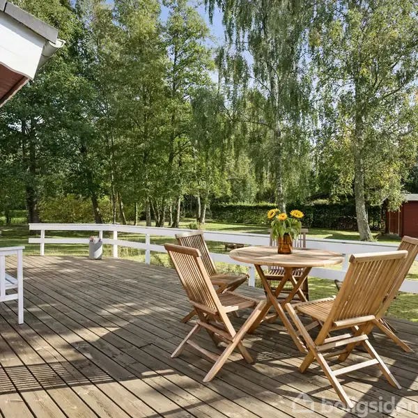 Et rundt havebord med tre træstole står på en terrasse, omkranset af et gelænder. En vase med solguler står på bordet. Omkring er der en grøn have og træer.