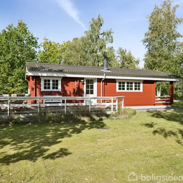 Rødt træhus står stille omgivet af frodig grøn have med høje træer. Huset har en veranda foran, og græsplænen er stor og solrig.
