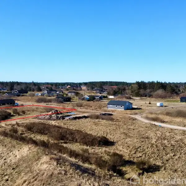 En grå bygning står på et åbent, græsklædt område omgivet af små huse i en landsby. En grusvej løber forbi, og røde linjer markerer områder på jorden.