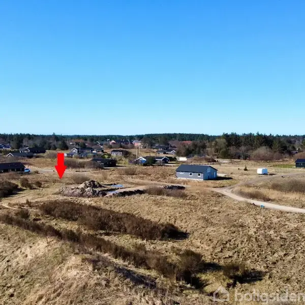 En mark med spredte huse i det fjerne. En rød pil peger ned mod et specifikt område. Det er en klar solskinsdag med en blå himmel.
