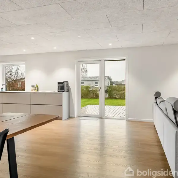 Køkken-alrum med spisebord og stol står på lyst trægulv. En grå sofa er i højre side. Store vinduer åbner ud til en grøn have udenfor.