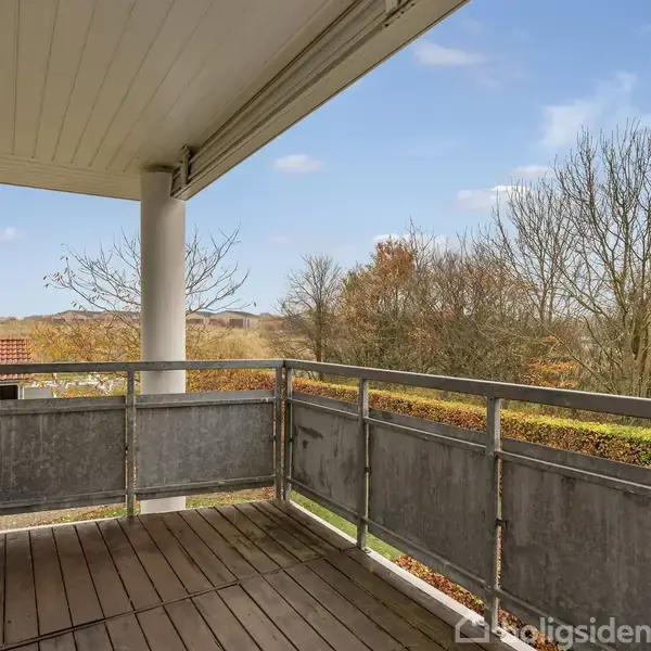 En træterrasse er tom og omgivet af et metalgelænder. I baggrunden ses træer uden blade og et rødt tegltag, med en klar blå himmel over. Ingen tekst til stede.