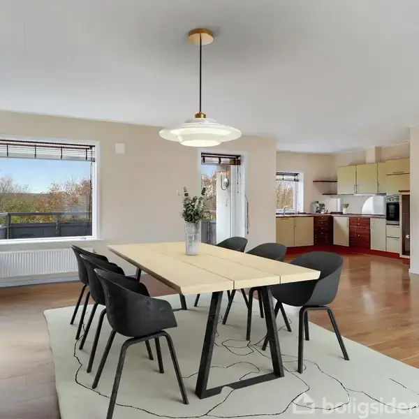Et spisebord med seks sorte stole står i et lyst, rummeligt rum med trægulv. I baggrunden ses et åbent køkken med lyse skabe. Vinduer giver udsigt til træer udenfor.