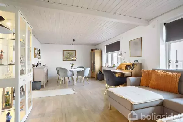 Stue med en grå sofa med orange puder, der står ved et skrivebord i hjørnet. Spisebord med stole og vitrineskab i hjørnet. Lys fra vinduer oplyser rummet.