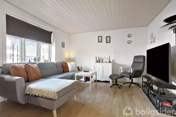 Grå sofa med orange puder står i stuen. Nær sofabordet er et sort tv. Vægge har billeder og lyse dekorationer, mens et vindue slipper lys ind. Lys trægulv.