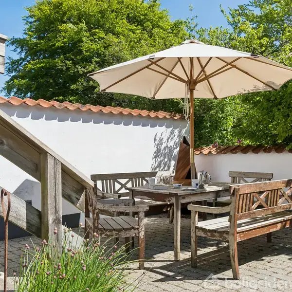 En træbænk og bord med parasol står på en stenbelagt terrasse. Omgivet af grønne hække og en hvid mur med teglkant.