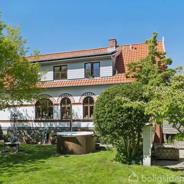 Hvidt hus med rødt tegltag står i en frodig have. Haven har et boblebad og grønne træer omkring. Klar blå himmel er synlig over huset.