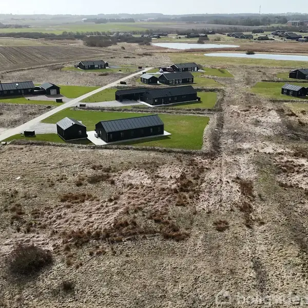 Flere sorte huse står spredt over et åbent landskab med grusveje og grønne plæner. Omkring dem er der marker og nogle små søer i baggrunden.