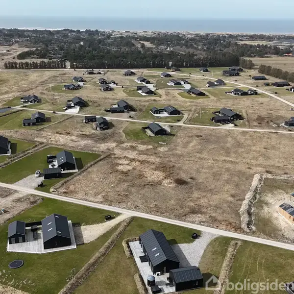 Flere moderne huse står spredt ud over et åbent, grønt landskab med grusveje, trægrupper og en sø i det fjerne.