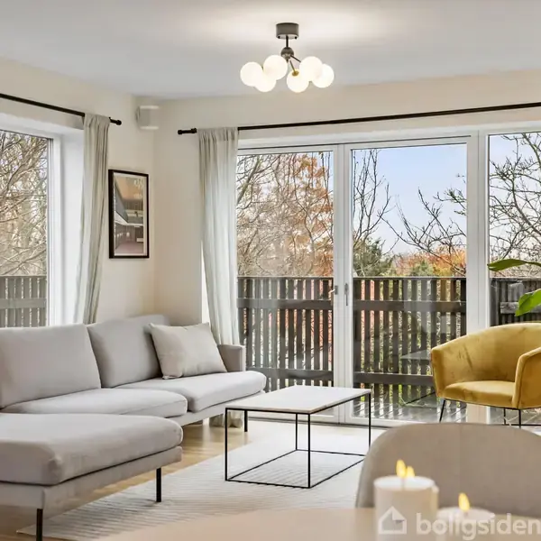 En lys stue med en grå hjørnesofa står ved siden af store vinduer med udsigt til en træterrasse. Et lille bord og en gul lænestol supplerer rummet.