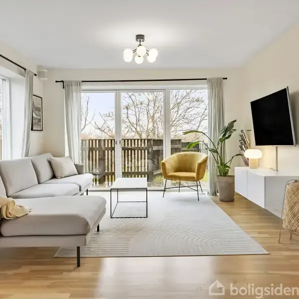 Stor sofa står i en stue med lyse træ-gulve. Et tæppe ligger foran sofaen. Et tv er monteret på væggen. Glasdøre åbner til en have med træer.