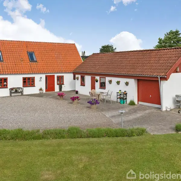 Et hvidt hus med rødt tegltag står i en velplejet have. Der er grus i gårdspladsen, og blomsterkrukker pynter indgangen. Garage og udendørs siddeplads er til stede.