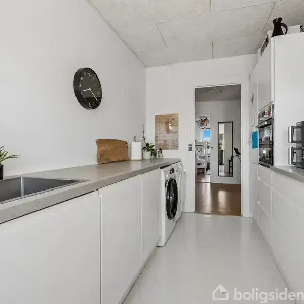 Et moderne køkken med hvide skabe, en vask ved siden af en grøn plante, og en vaskemaskine under bordet. En mikroovn og ovn er indbygget til højre. Væggen har et rundt ur.