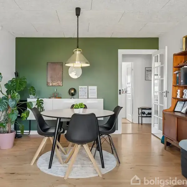 En spisebord med sorte stole står i en lys spisestue. En grøn væg bagved er dekoreret med kunstværker. En reol med bøger og nipsting står til højre.