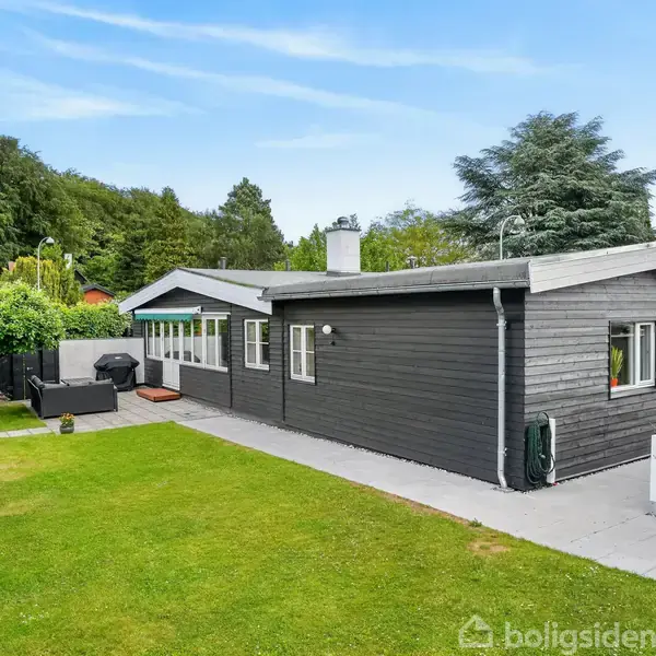 Et sortmalet hus med fladt tag står i en grøn have. Langs husets facade er der terrasse med havemøbler og grill. Haven er omgivet af høje træer og buske.