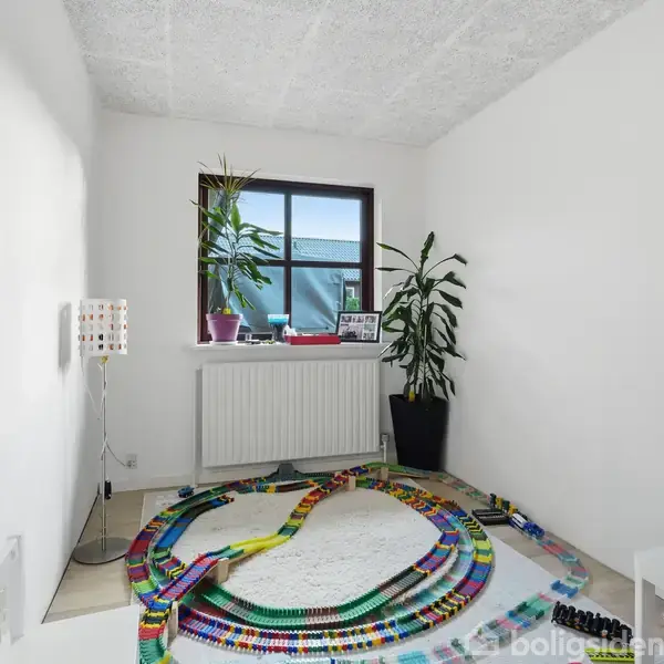 Et rum med en modeljernbane, farvestrålende og snoet på gulvet. To store planter står ved et vindue. En lampe er monteret på væggen, og der er en radiator under vinduet.