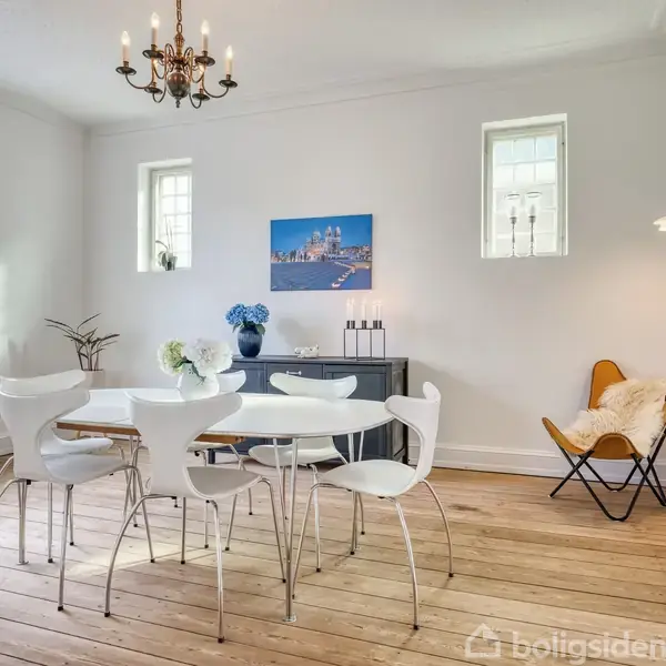 Et lyst spisestueområde med et rundt bord, omgivet af hvide stole. På væggen hænger et maleri. Trægulv og en lysekrone bidrager til den elegante atmosfære.