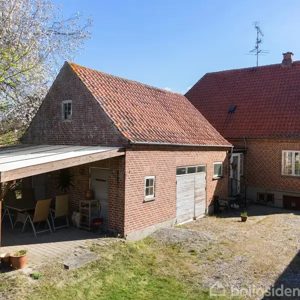 Et rødt murstenshus med rødt tegltag står i en grøn have. Ved siden af er en garage med en overdækket terrasse, hvor der står havemøbler.