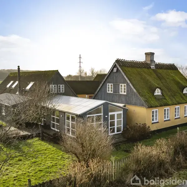 Et hus med stråtag står stille på en grøn mark. Himlen er blå med spredte skyer, og der er træer omkring huset.