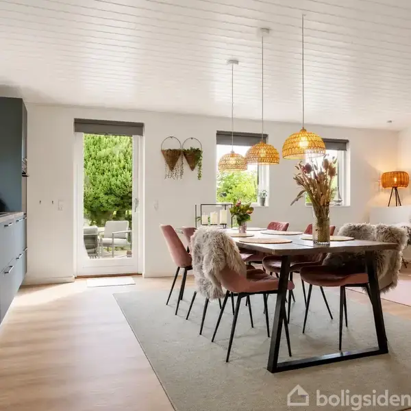 Spisebord omgivet af stole med pelsbetræk står i moderne, lys stue. Væglamper og store vinduer mod terrasse skaber en indbydende atmosfære. Planter og kunstværker dekorerer rummet.