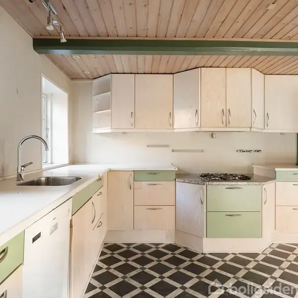 Køkken med hvide og grønne skabe, vask og gaskomfur, midt i et rum med træloft og sort-hvidternet gulv. Enkelt vindue ved siden af vasken lyser rummet op.