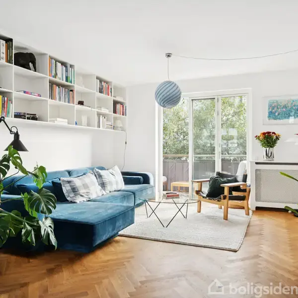 Blå sofa står foran en hvid reol fyldt med bøger. Stuen er lys og moderne med trægulv, store vinduer og planter. En lampe hænger over sofabordet.