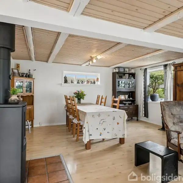 Stue med brændeovn til venstre, spisebord med seks stole i midten, hyggekrog med lænestol til højre. Lyse trælofter og parketgulv skaber en varm, indbydende atmosfære.
